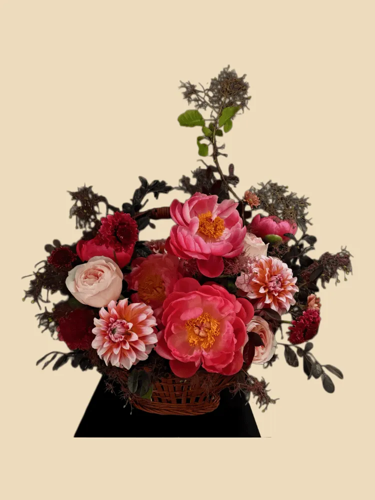 Full coral peonies, blush garden roses, pink-orange dahlias, scabiosa, dark carnations, and burgundy foliage in a basket