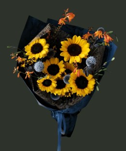 Full dark-centred yellow sunflowers with steel-blue eryngium, orange crocosmia, small daisies, and dark foliage in navy wrapping
