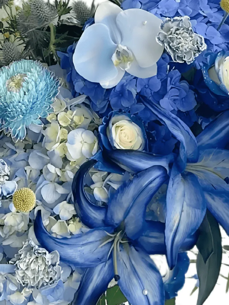Close-up of dyed cobalt lilies, blue hydrangea, marbled roses, pale orchids, chrysanthemums, eryngium, and yellow craspedia in porcelain